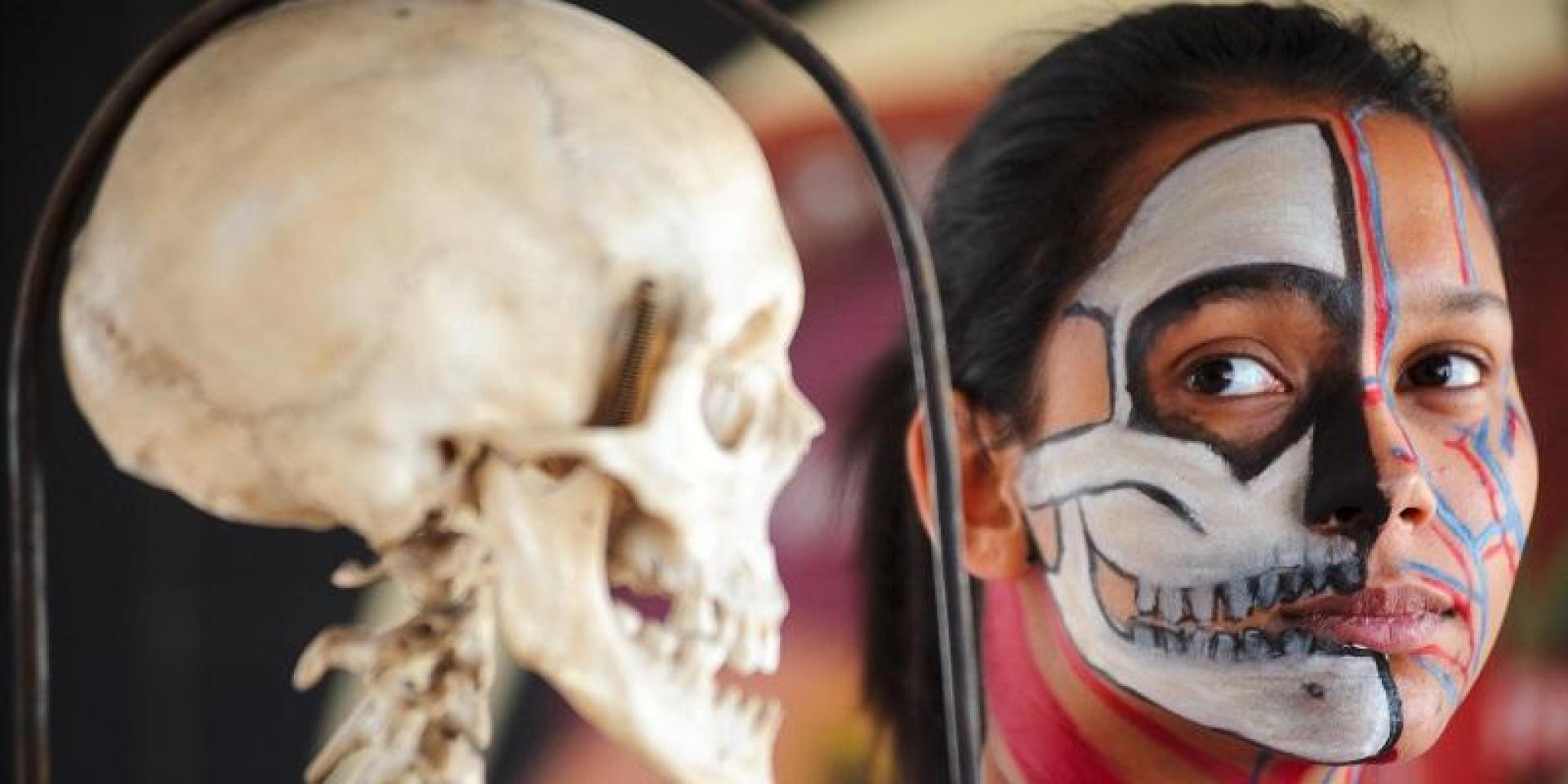 A girl with her face painted with the image of a skull next to a skull