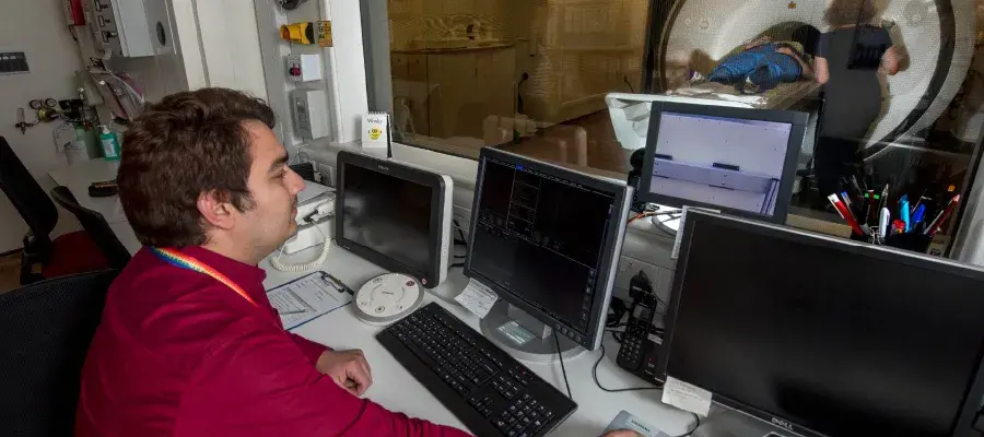 Man operating medical imaging equipment from behind a pane of glass