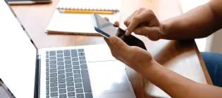 Close up of hands checking a mobile phone with laptop in background
