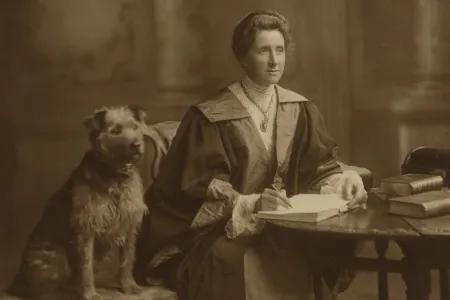 Black and white image of Jessie, in graduation robes, sitting at a desk with a dog at her side