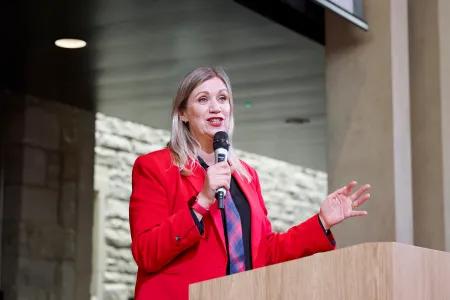 Marti Balaam in red jacket holding microphone and speaking at a podium