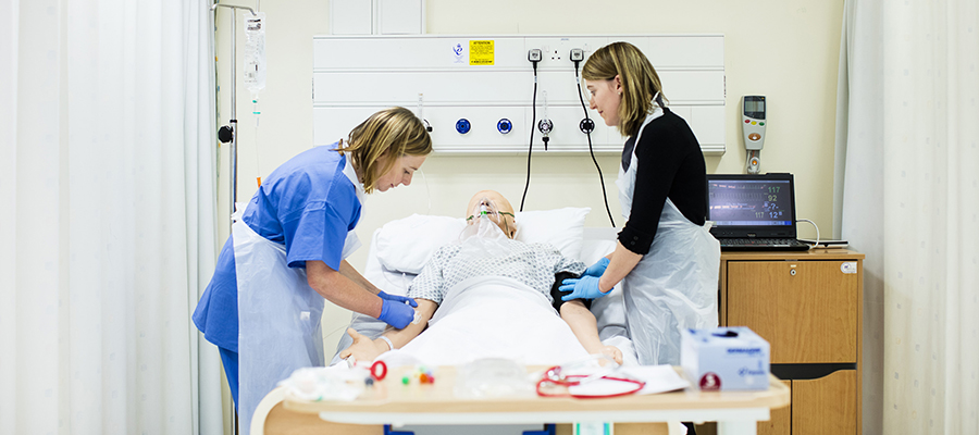 Two members of the Clinical Skills Centre staff stand over a SimMan mannequin in a makeshift hospital bed